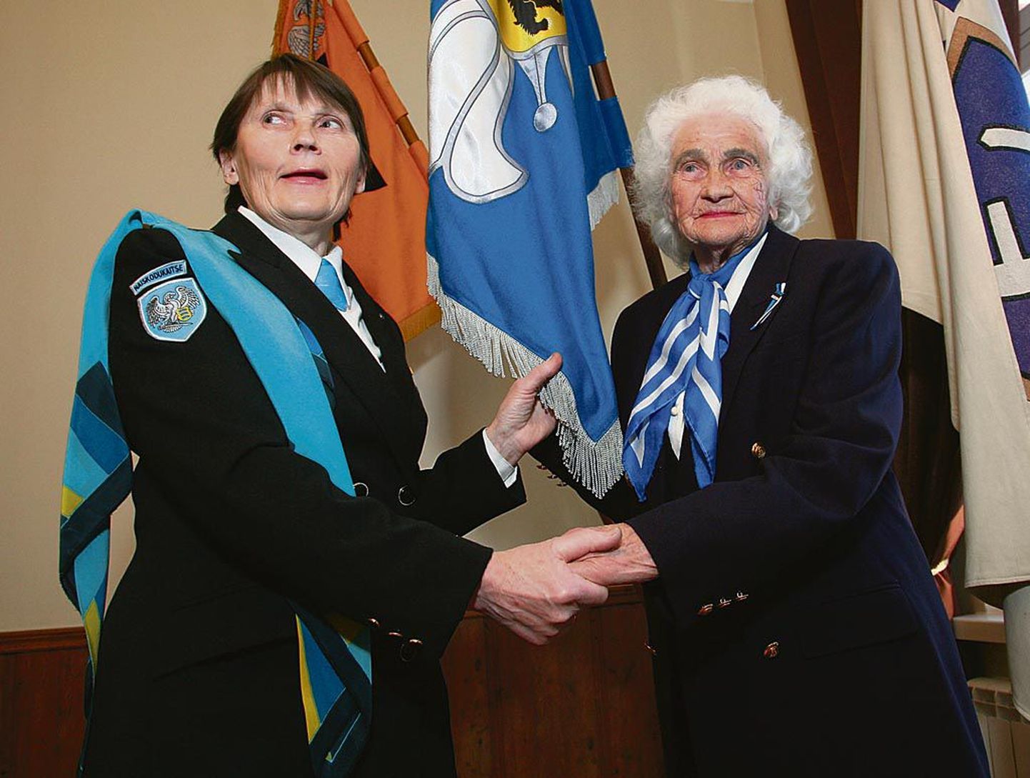 Naiskodukaitse Pärnumaa ringkonna auliige Elfriede Laidvee ja juhatuse liige Maie Pitsal on ulatanud teineteisele käe ringkonna lipu ees.