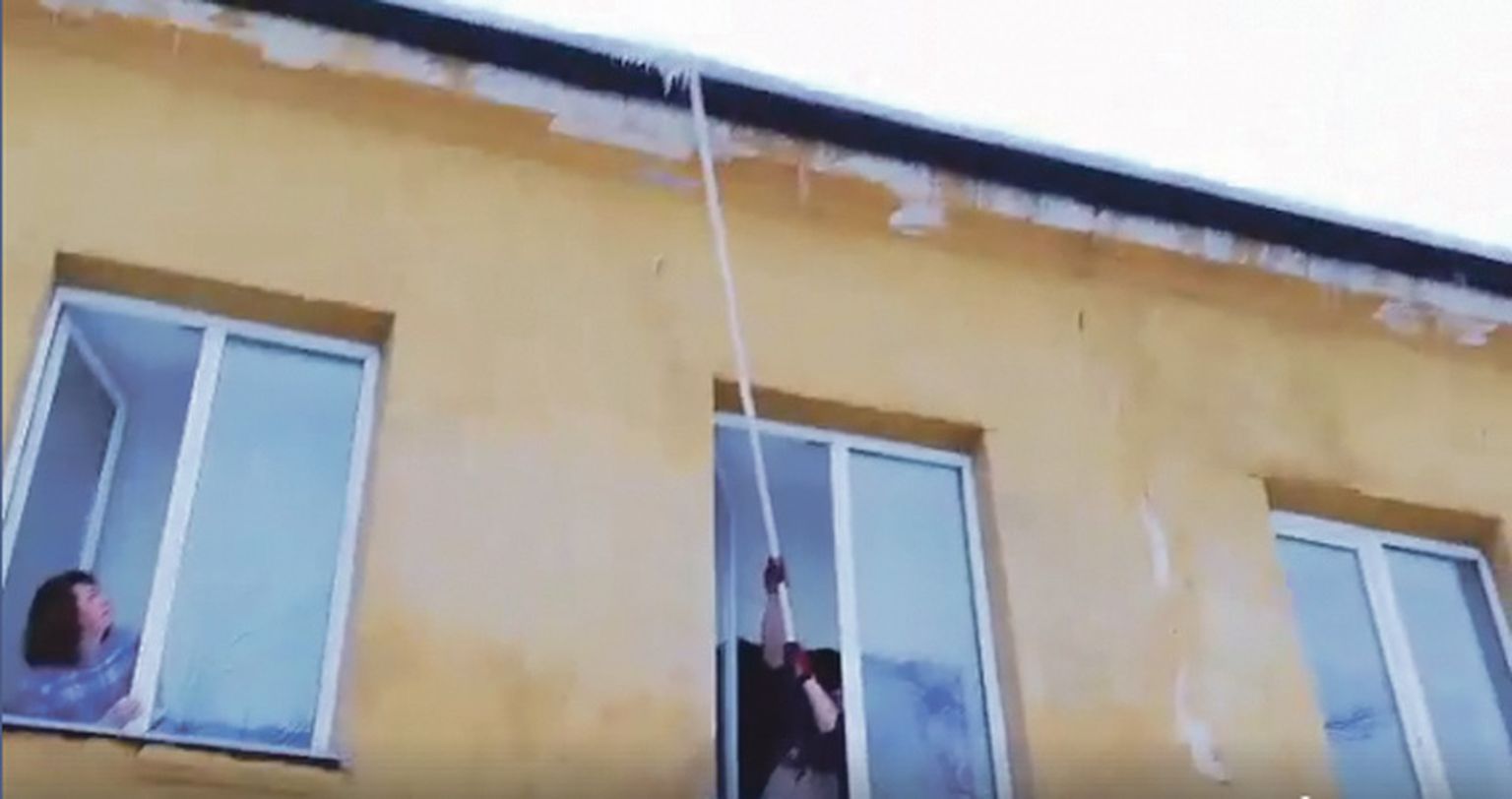 Кадр из видео, на котором видно, как работник, стоя у окна, пытается сбить длинной палкой сосульки с края крыши. Из соседнего окна за его действиями наблюдает директор школы Юлия Каламаева.