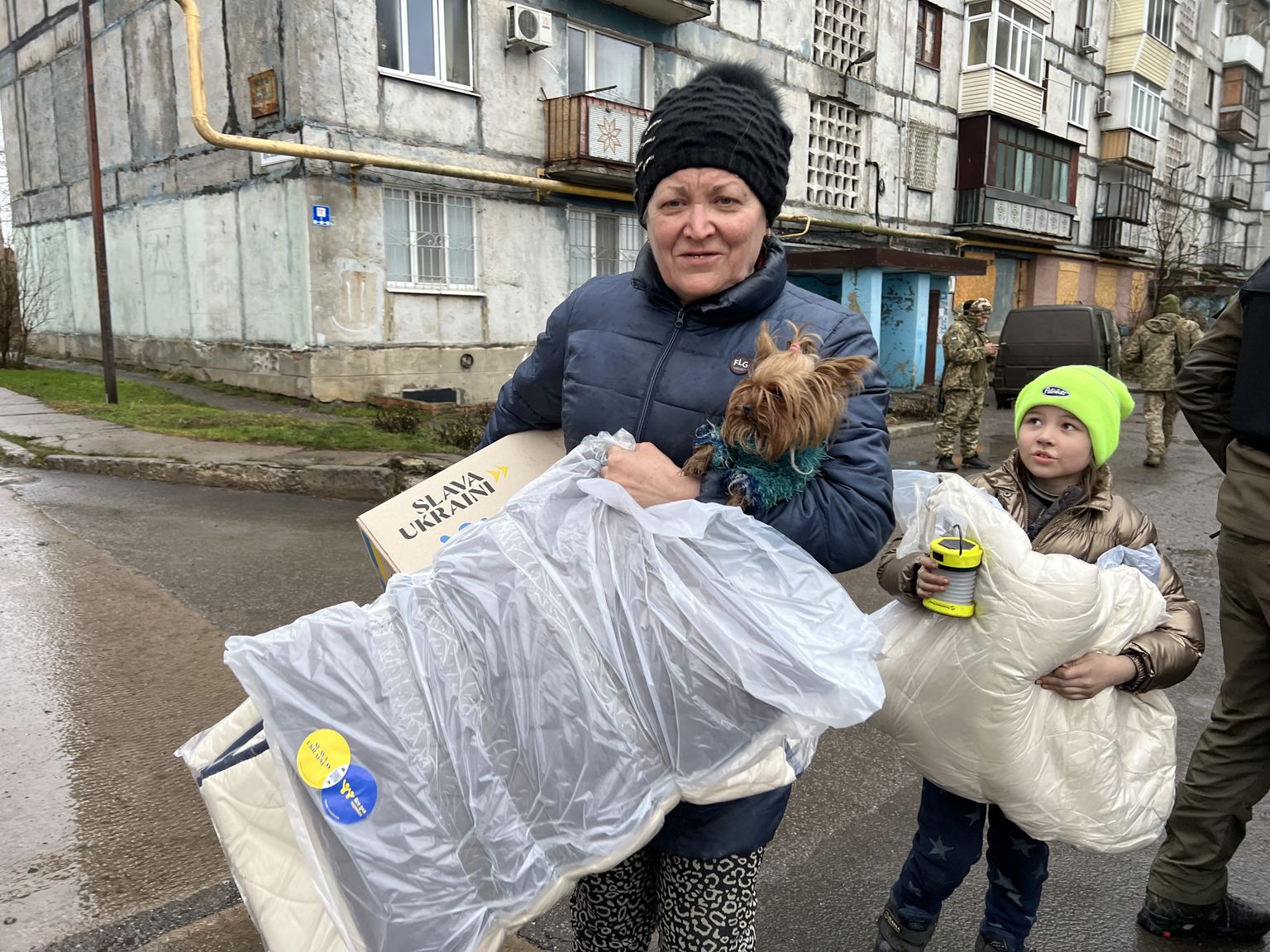 Vanaema Ljubov ja lapselaps Ljubov saavad Bahmutis Eesti MTÜ Slava Ukraini toodud humanitaarabi - kasti toiduainete, hügieenitarvete ja taskulambi ning küünaldega, samuti sooja teki. Nende taga on näha Ukraina sõjaväelased, kes valmistuvad lahinguteks linnas.