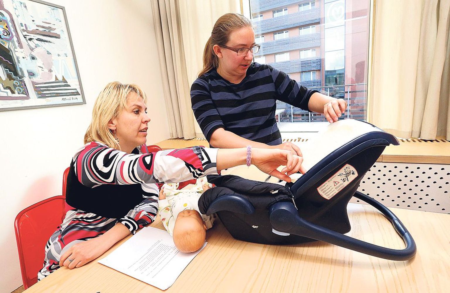 Õppejõud Kristi Vahur (vasakul) ja Tartu tervishoiukõrgkoolis tervisekaitse spetsialisti eriala omandanud Elen Pabo näitavad, miks vana turvahäll beebile autovarustuseks enam ei sobi. Laual lebab nukk, mitte laps.