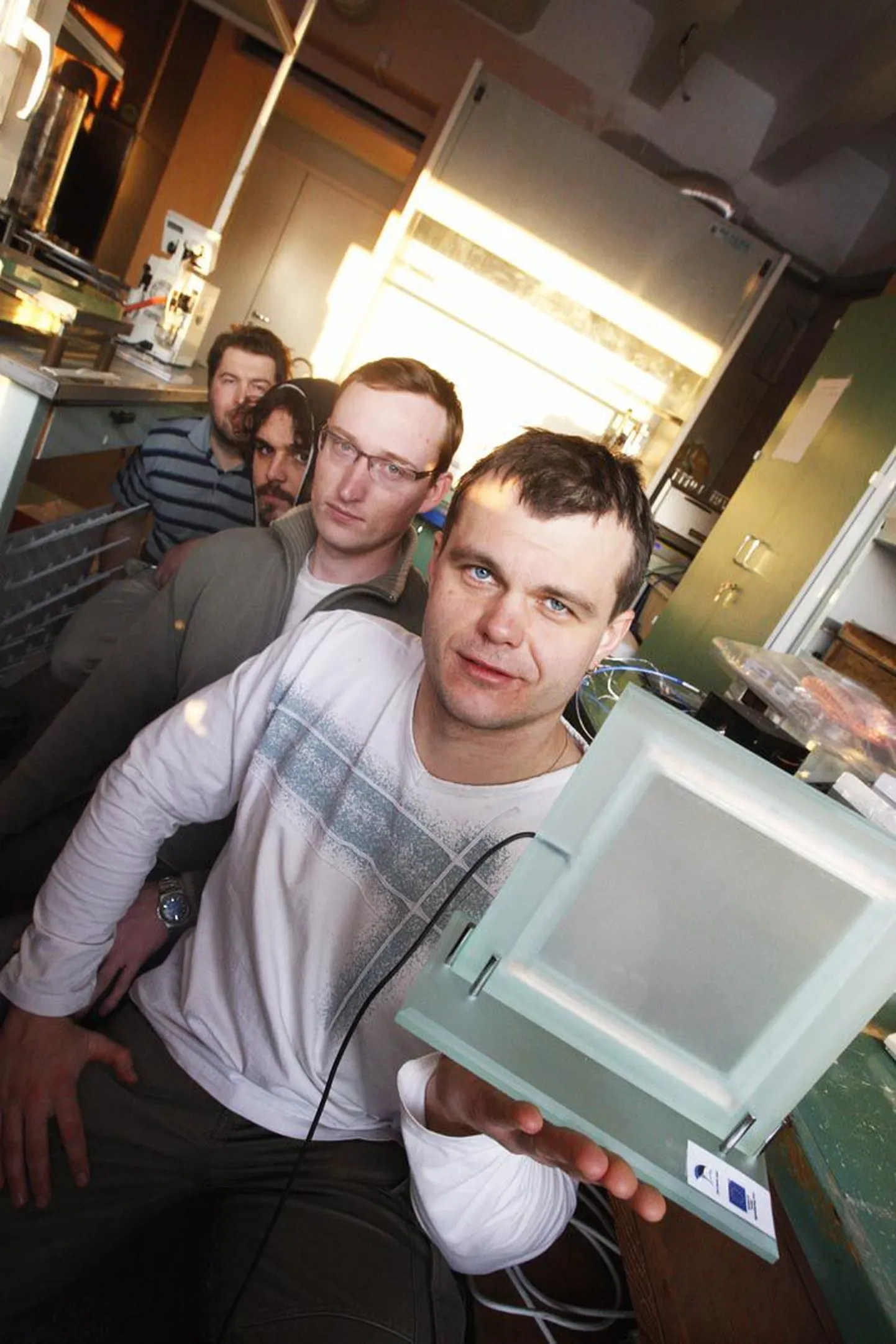 Teadlased Kristjan Saal (esiplaanilt), Martin Timusk, Martin Järvekülg ja Rünno Lõhmus näitavad oma laboratooriumis juhtmetega ühendatud klaasitükki ehk elektrooptilist klaasi.