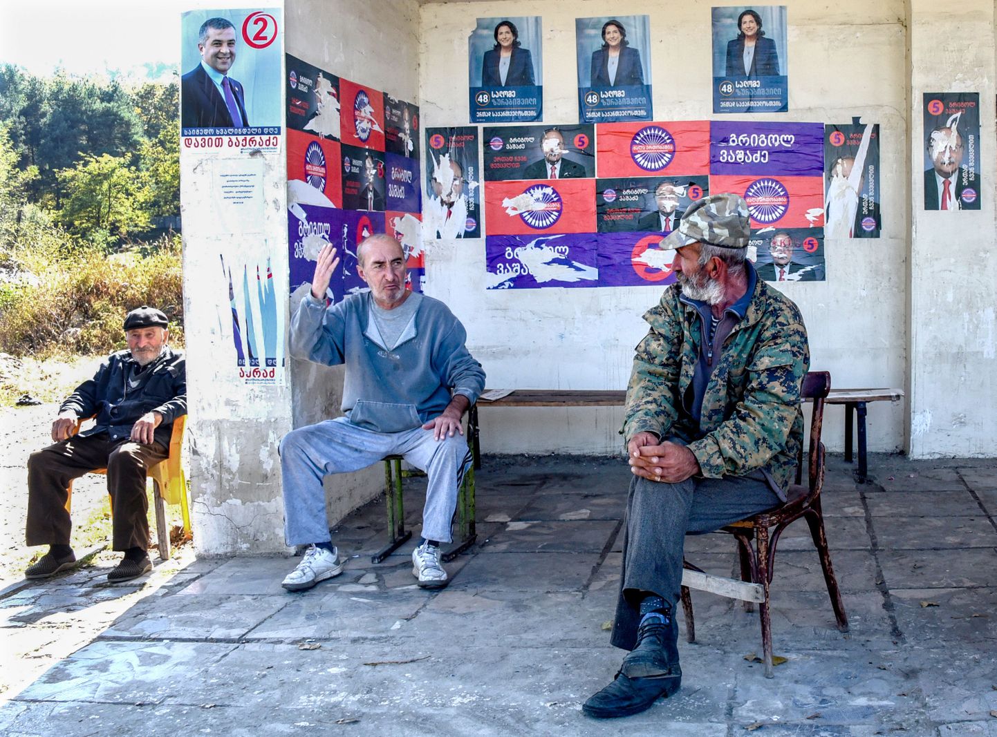 Vīrieši sēž ar vēlēšanu aģitācijas plakātiem aplīmētā autobusa pieturā Gruzijas pilsētā Telavi
