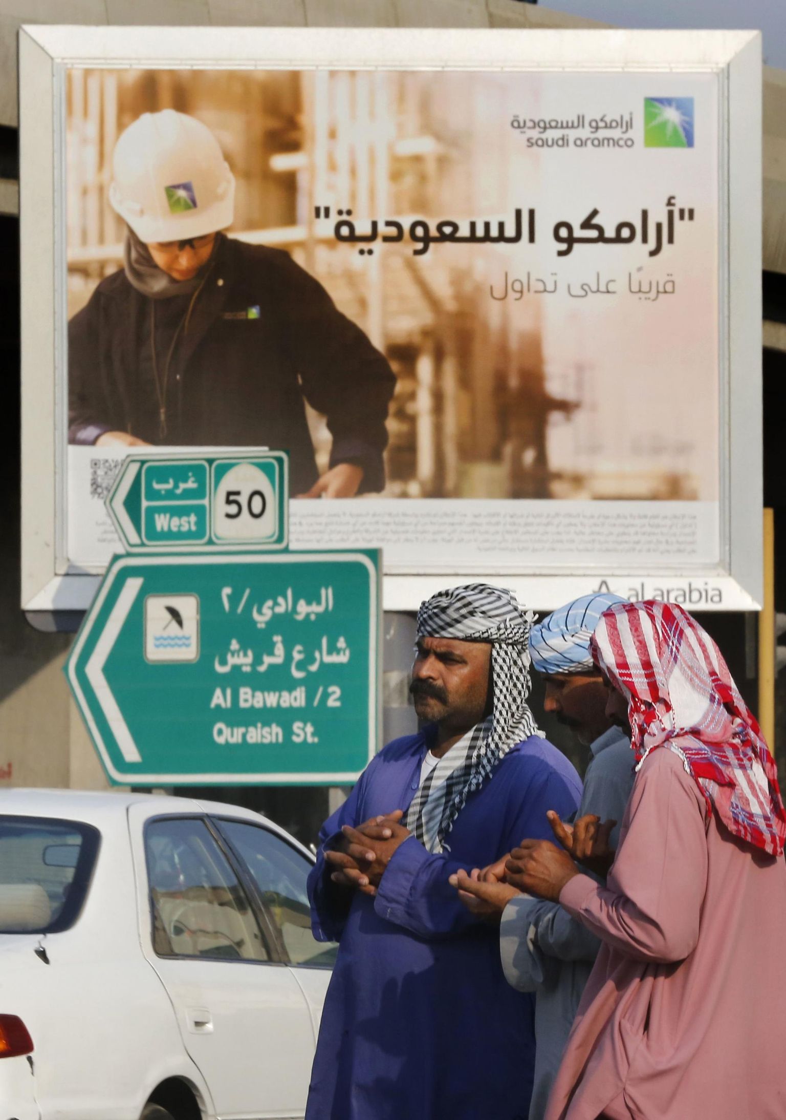 Kohalikud töötajad plakati ees, mis reklaamib Saudi Aramco aktsiamüüki: araabiakeelsel plakatil seisab: «Saudi Aramco, peagi börsil».