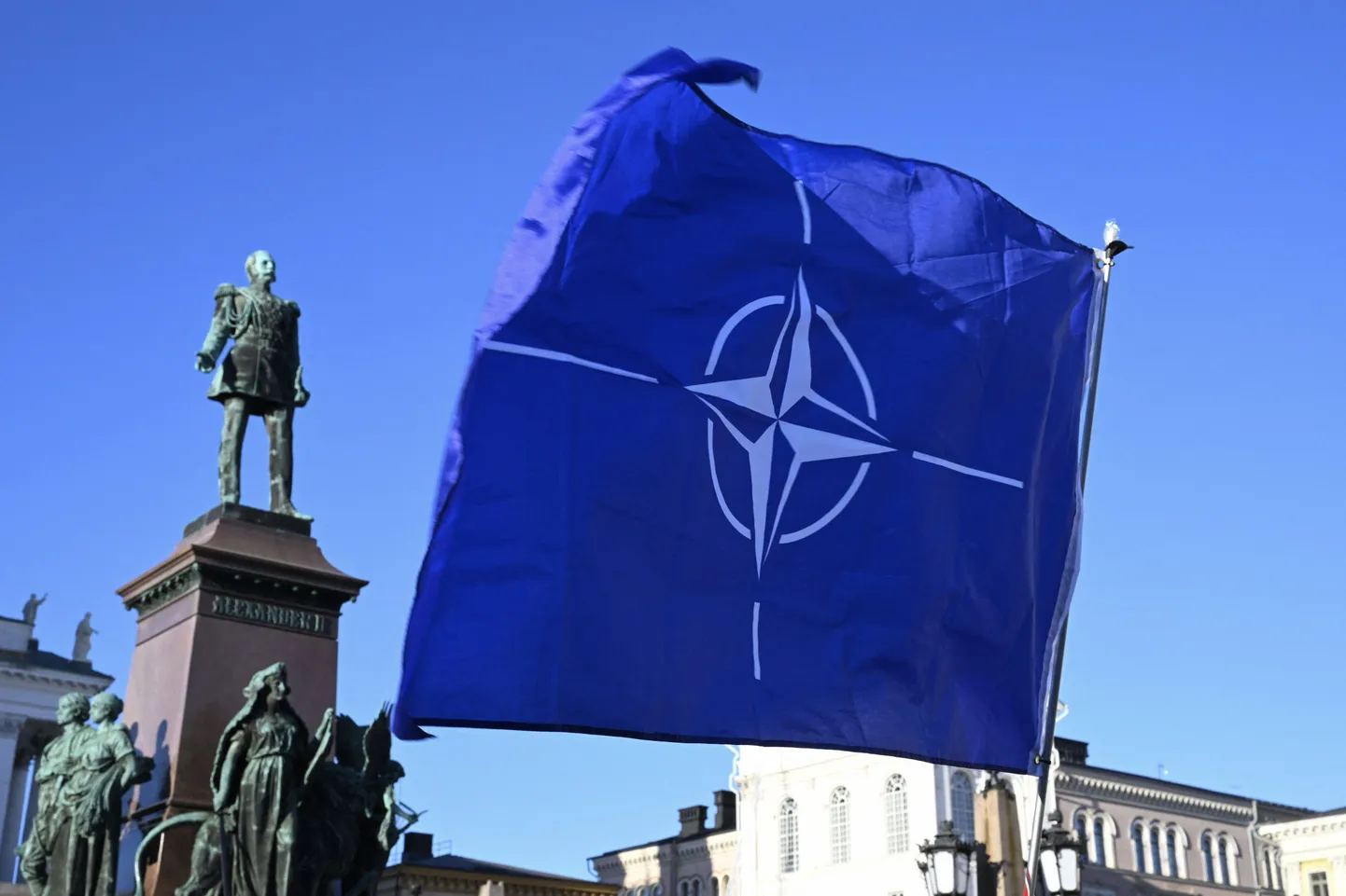 Флаг НАТО на Сенатской площади в Хельсинки в руках горожан, которые отмечают встпуление Финляндии в военный альянс, 4 апреля 2023 года. На фоне флага памятник великому князю Финляндскому и российскому императору Александру II.